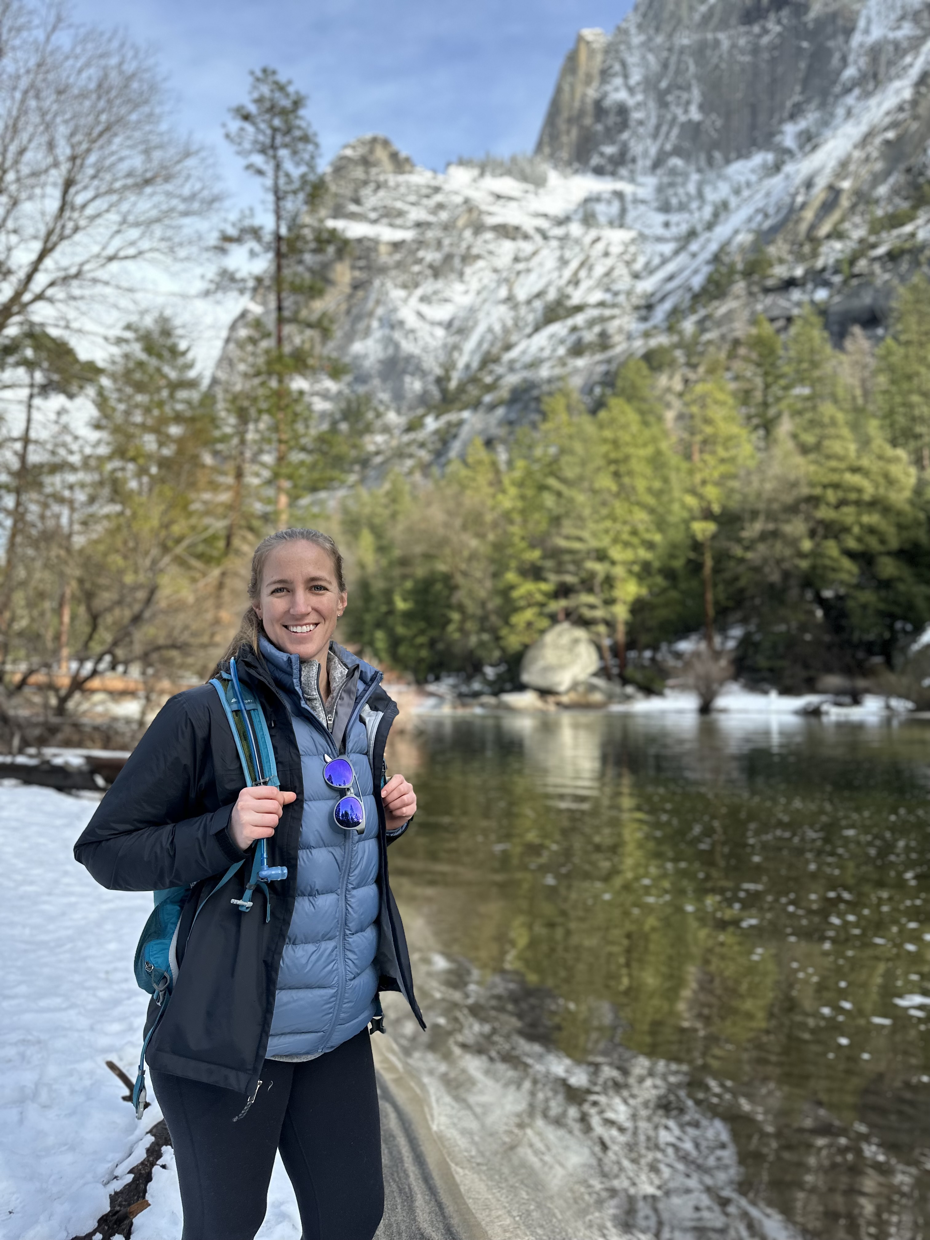 A woman wearing warm layers and a backpack poses next to a river with snow and mountains in the background