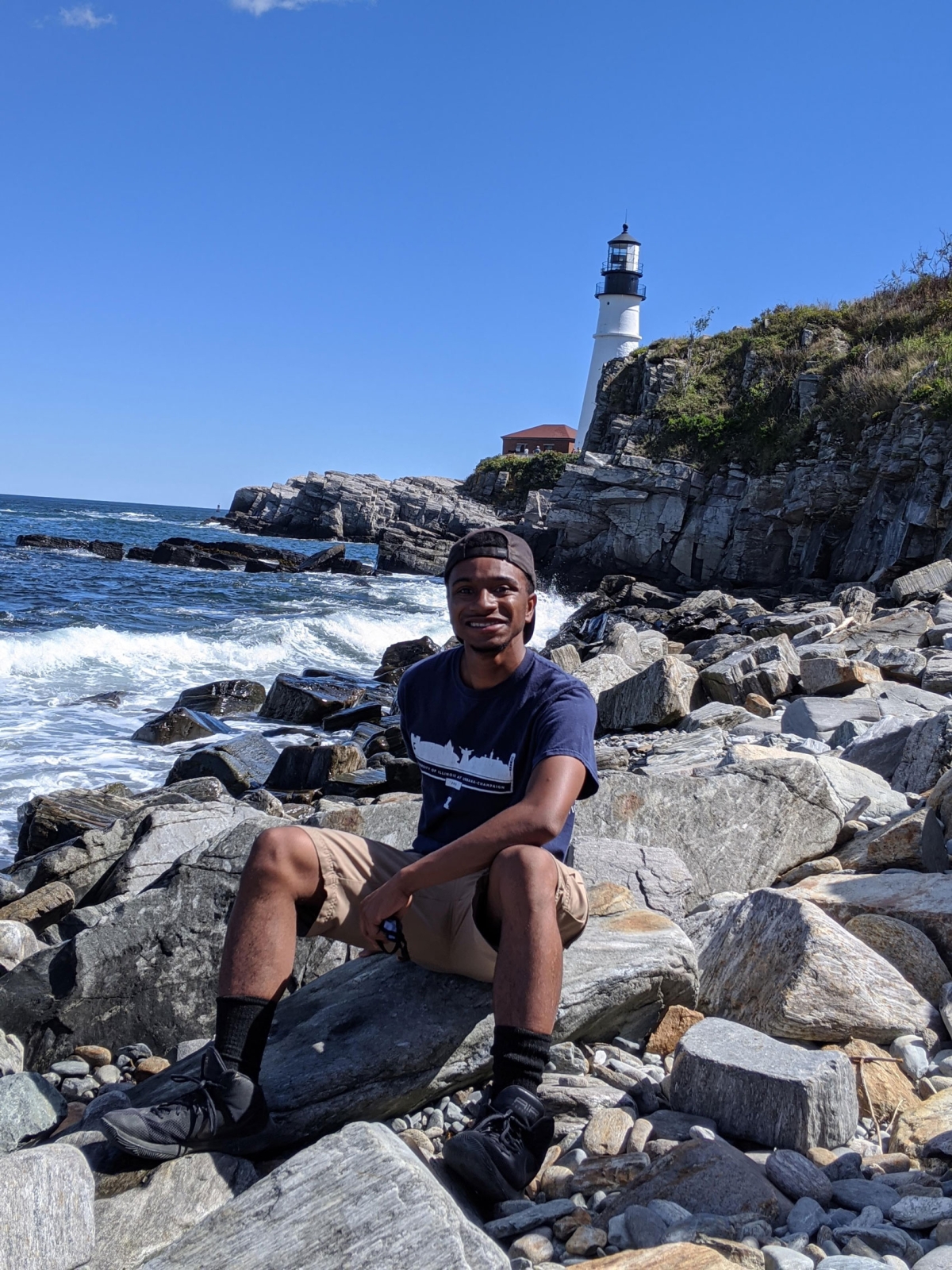 Kai Hardy sits on rocks near a body of water with a lighthouse in the background